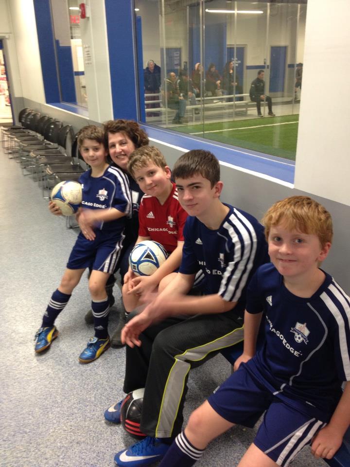 A mom poses with four children in sports uniforms.