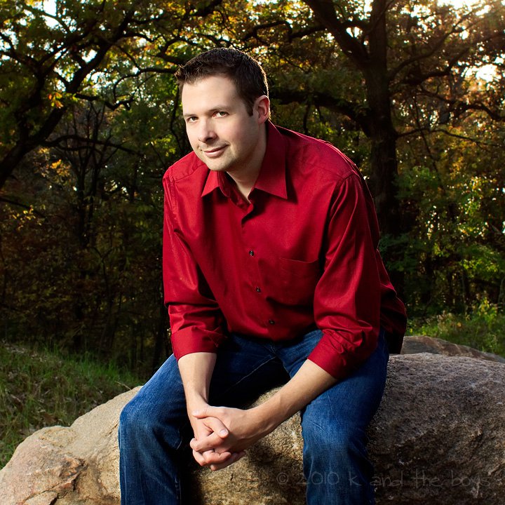 A man sits on a rock outside in front of some trees