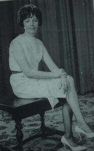 An old photo of a woman in the 50s or 60s, in a smart dress and heels, and sitting on a bench.