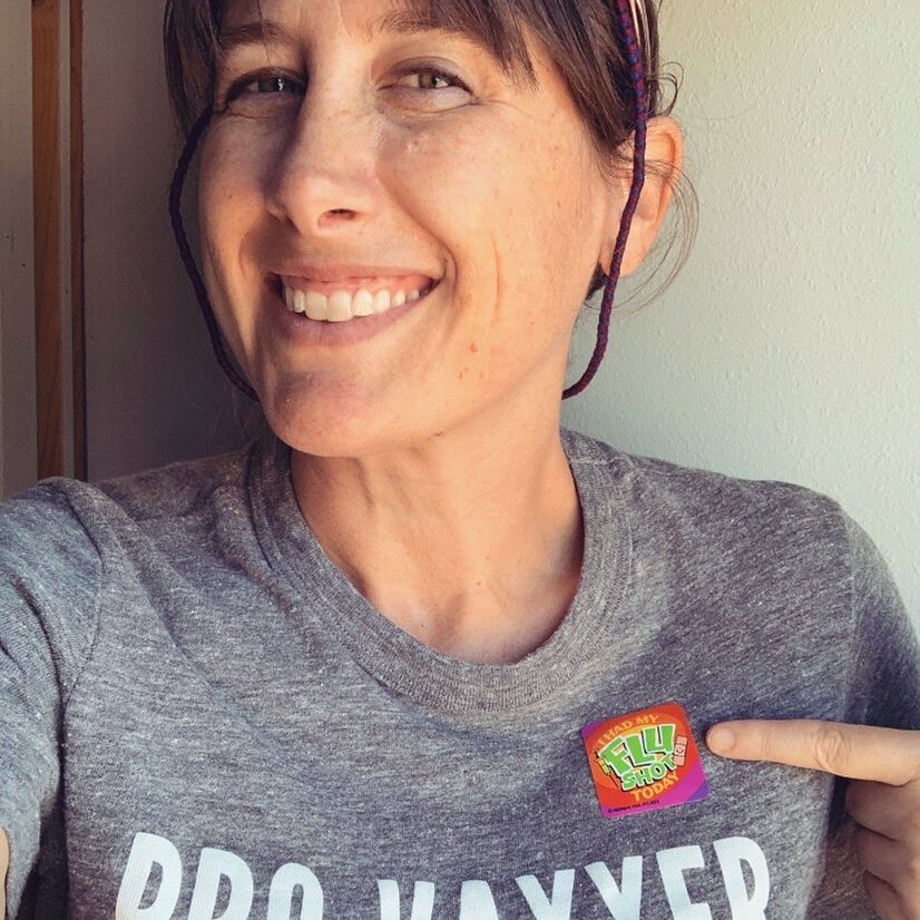 Lindsay Diamond wearing a grey "pro-vaxxer" shirt and pointing to a colorful sticker on that shirt.