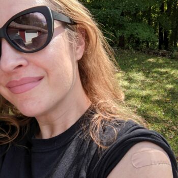 White Woman Wearing Sunglasses And Black Shirt With A Bandaid On Her Arm.