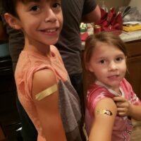 Two Small Children Proudly Show Off Their Vaccine Bandages.