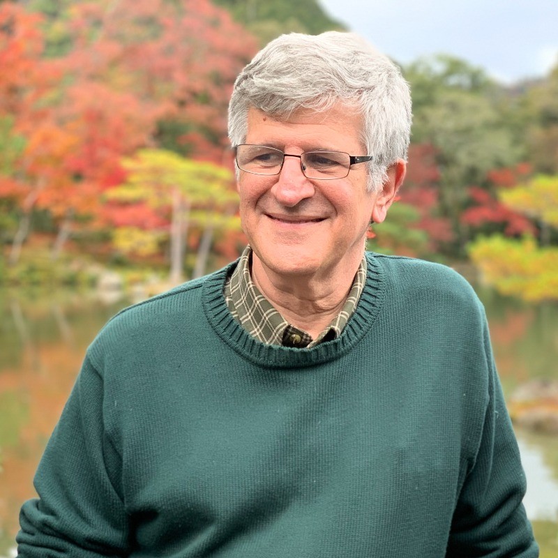 A photo of a bespectacled white man in a green sweater in front of a fall foliage.