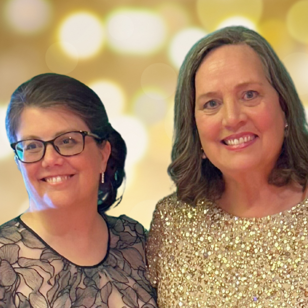 A bespectacled woman stands beside another brown-haired woman. Both are wearing fancy dresses.