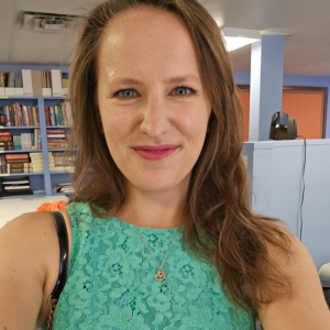A Woman In A Turquoise Lace Top Smiles While Taking A Selfie In A Room With Bookshelves In The Background.