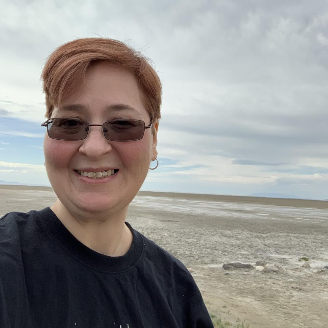 A selfie of Alysia with ginger hair and glasses, smiling in a black shirt against a desert landscape background.