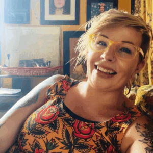 A woman with thick yellow glasses and a colorful floral shirt smiles at the camera.