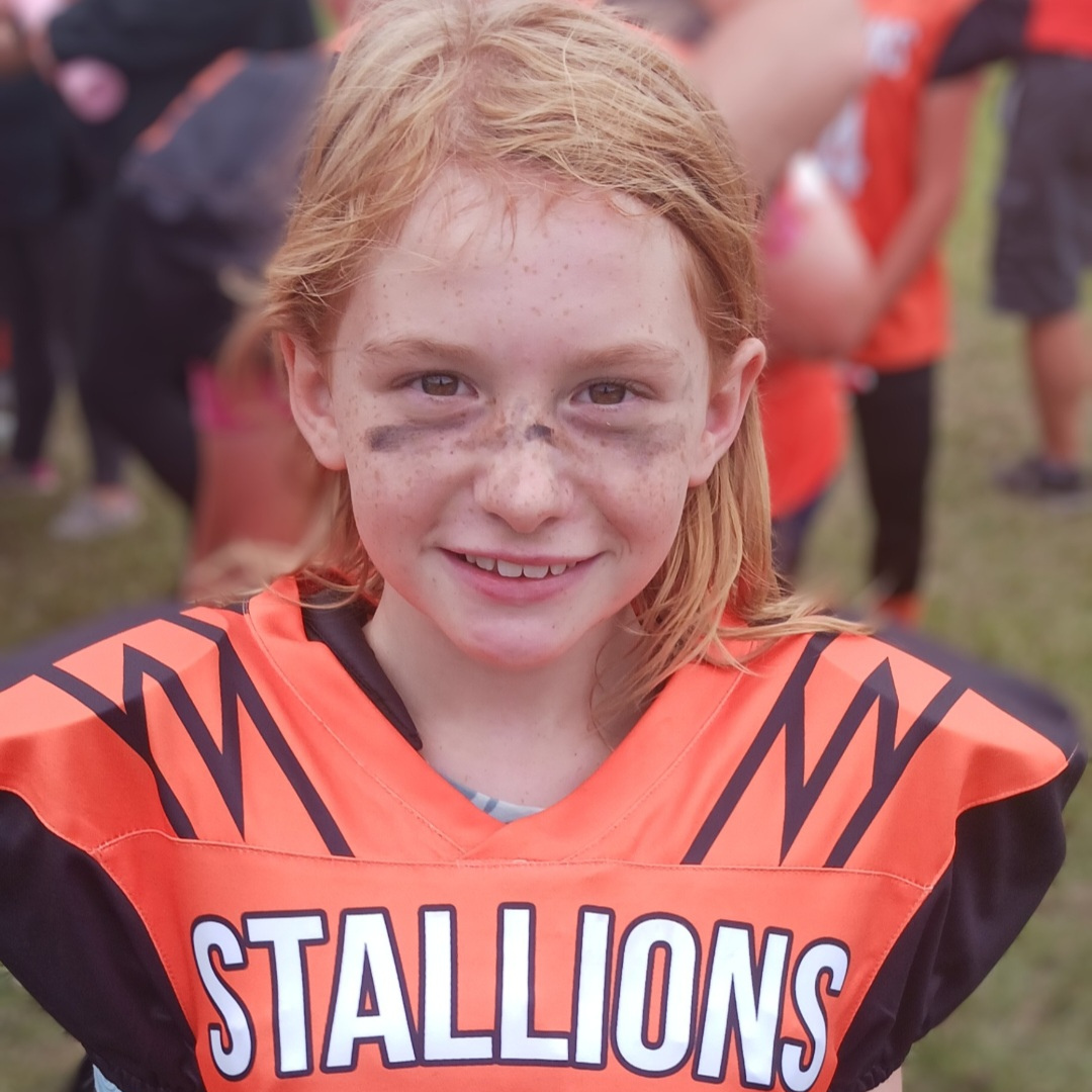 A young girl with orange hair in a dark orange football jersey that says "Stallions"