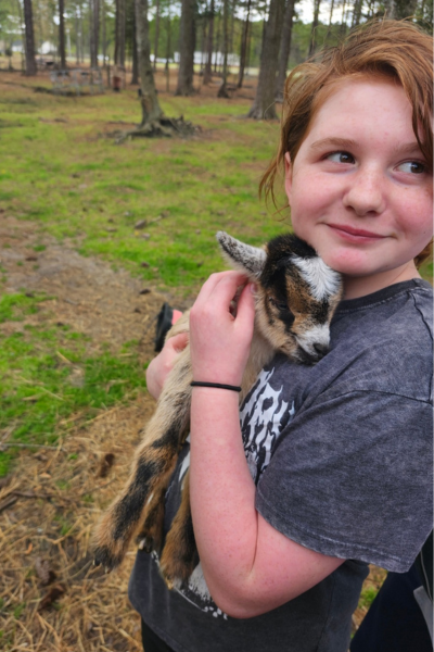Sophia stands in a forest holding a baby goat agaisnt her chest.