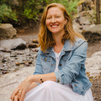 A woman in a jean jacket and white dress sits near a river.