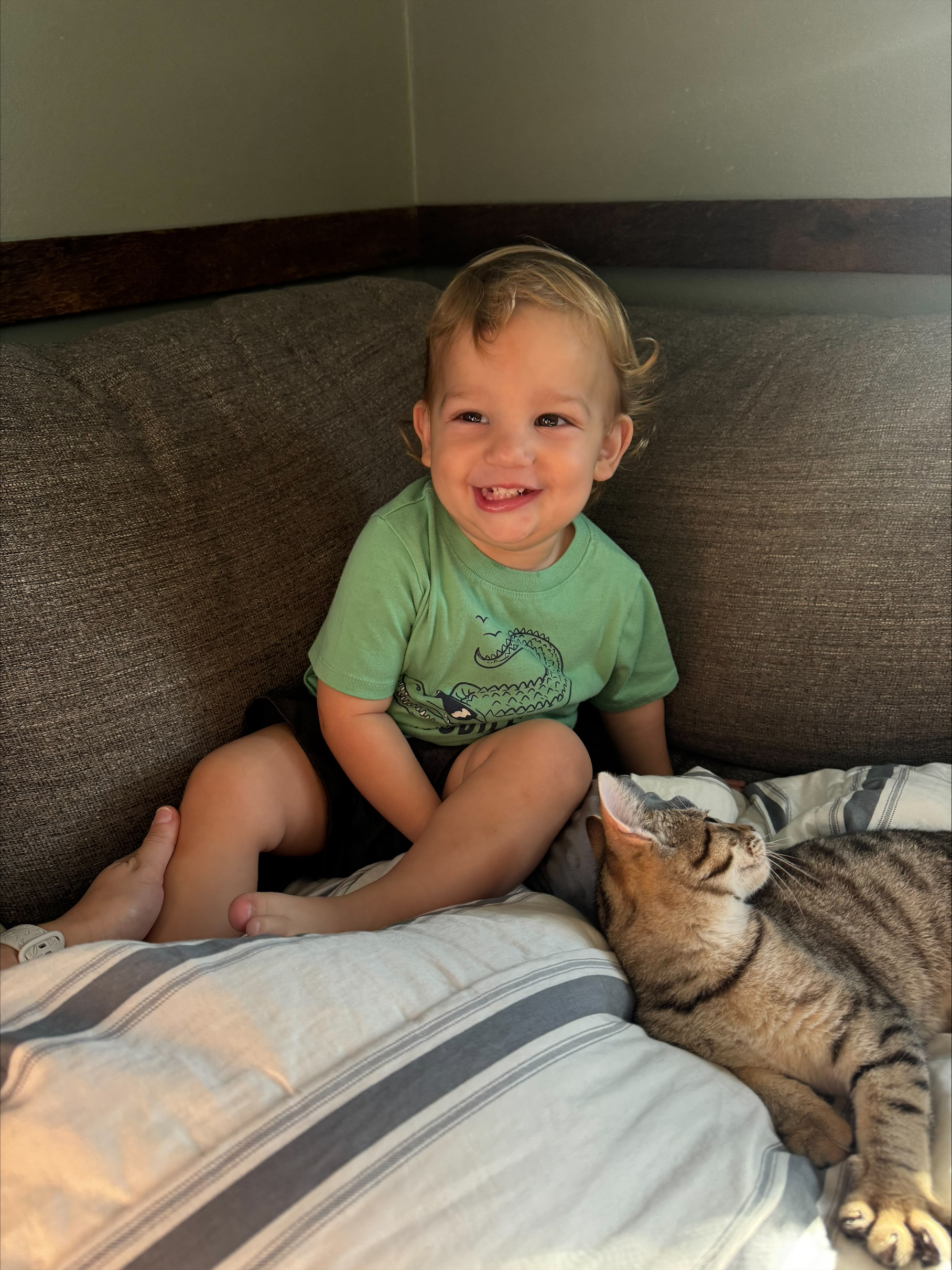 Toddler Logan grinning on a couch beside a sleeping tabby cat