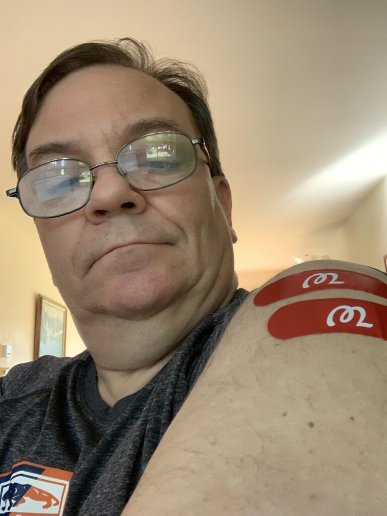 Man with wire-rimmed glasses and a gray T-shirt taking a close-up selfie, showing two red bandages on his shoulder.