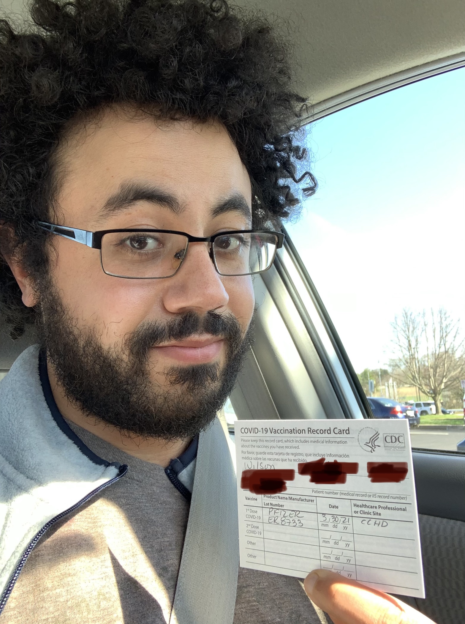 Dan Wilson, a man with curly hair and glasses holding a COVID-19 vaccination card while seated in a car.