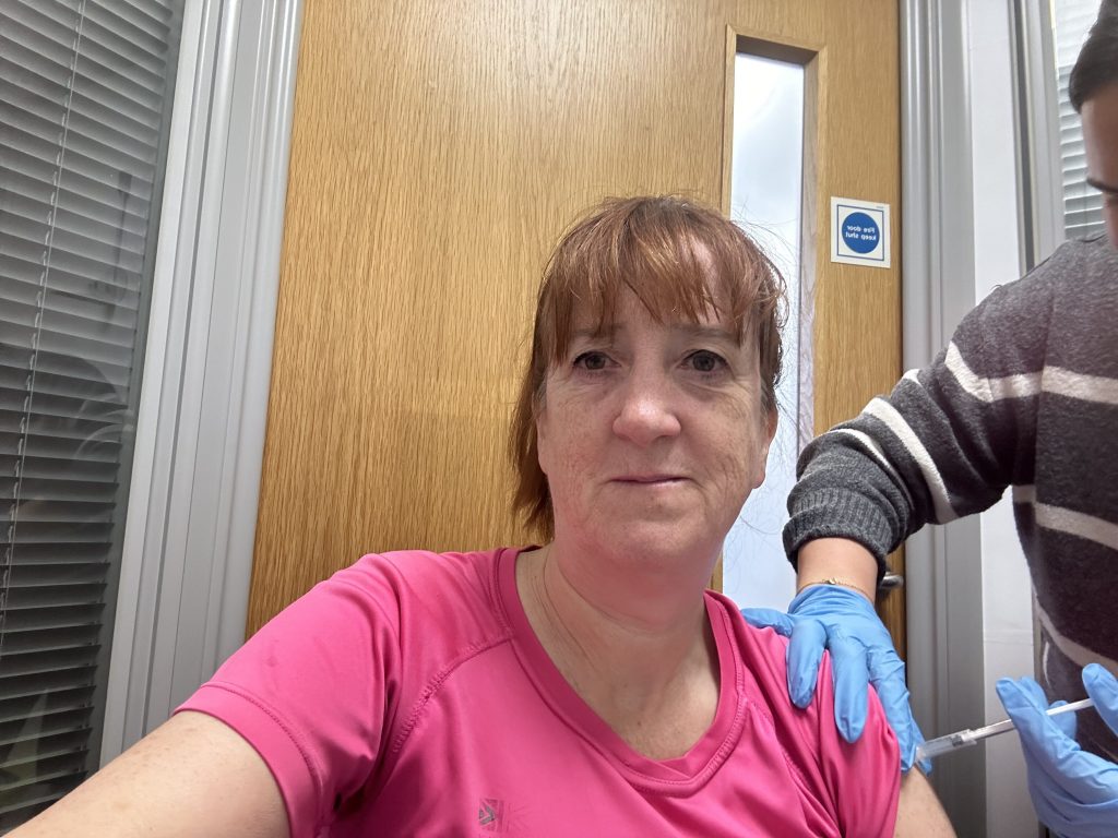 Woman in a pink top taking a selfie while a gloved worker gives her a shot.