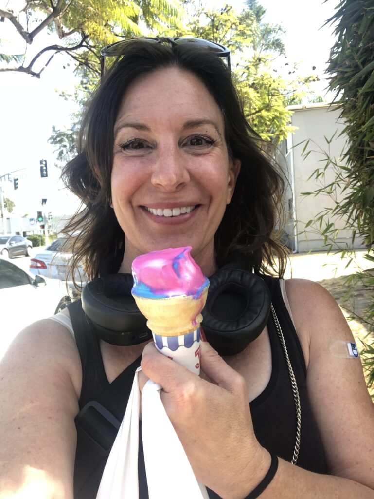 Smiling woman holding an ice cream cone outdoors, a small bandage on her upper arm.