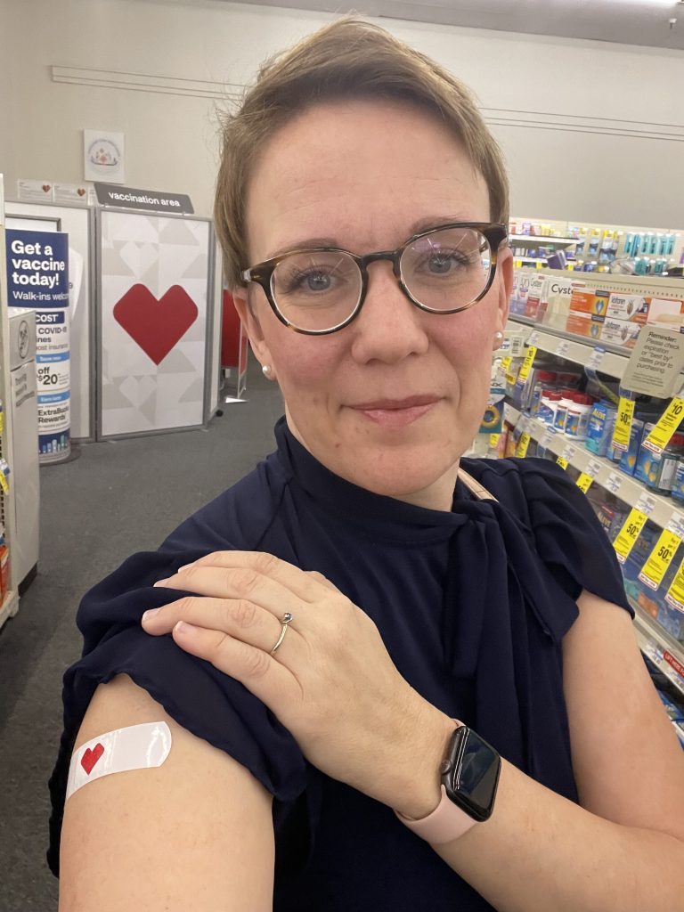 Woman in glasses in a pharmacy, her hand on her shoulder above a heart bandage on her upper arm.