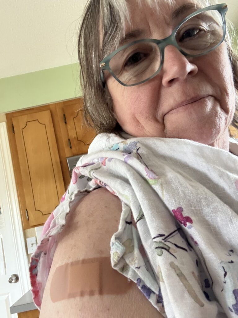Older woman in glasses showing two bandages on her upper arm in a kitchen.