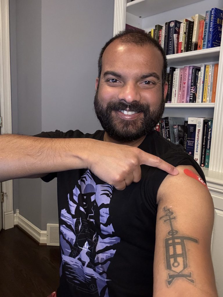 Man pointing at a red bandage on his upper arm, bookshelves behind him.