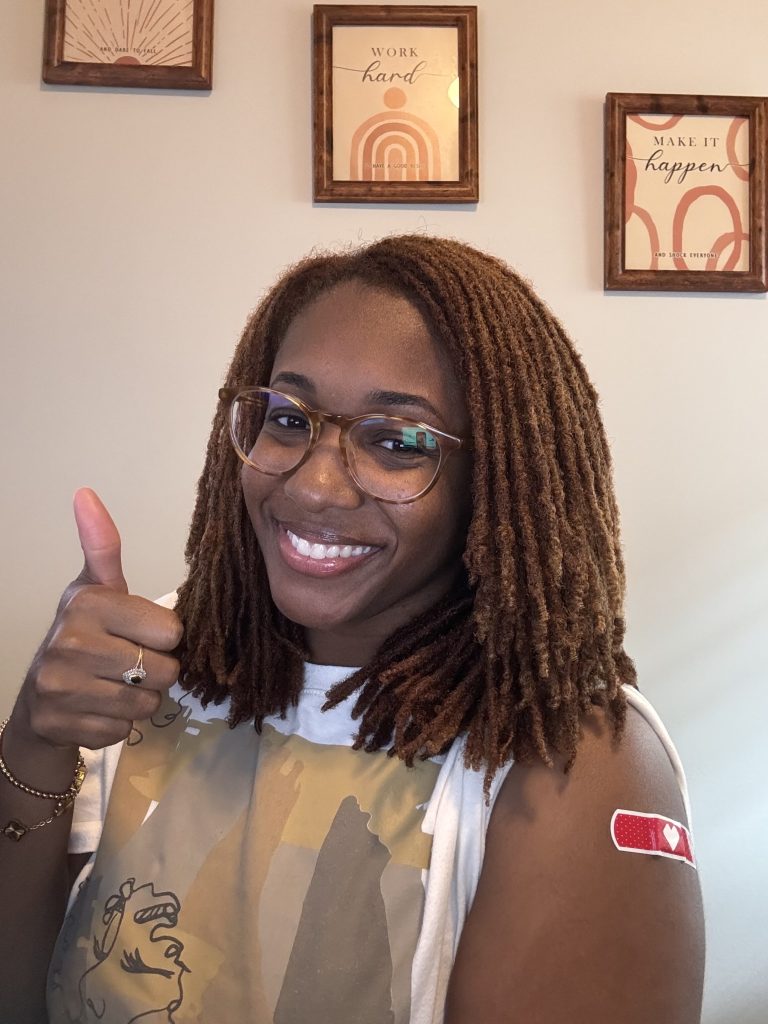 Smiling woman with locs and glasses giving a thumbs up, a red bandage on her upper arm.